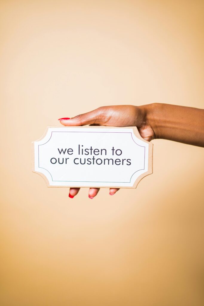 pexels-photo-7564196-7564196 A hand holding a sign saying 'we listen to our customers' on a neutral background.