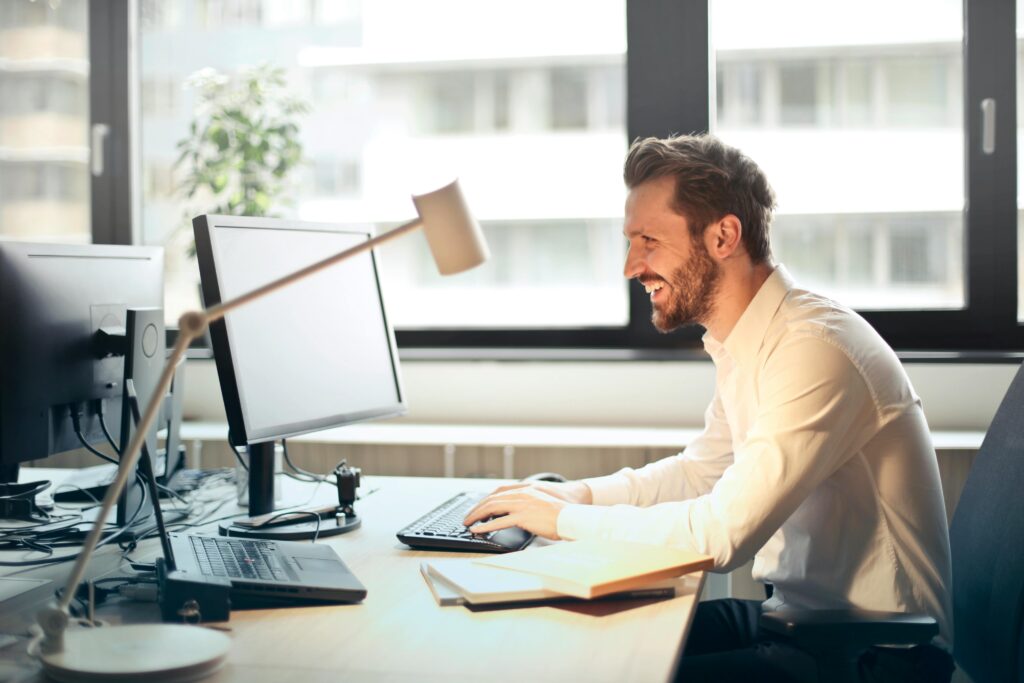 pexels-photo-840996-840996-1 A man smiling while working at an office desk with a computer and natural daylight streaming in through large windows.